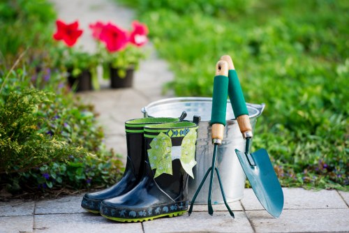 A garden scene in Gipsy Hill featuring a paved pathway with a pair of black rubber gardening boots with a floral pattern, placed alongside a white plastic bucket, hand tools including a small trowel with a blue handle and a garden fork with wooden handles, all set against a lush green background with blurred pink flowers and plants. The image captures outdoor gardening equipment arranged neatly, suggesting preparation for planting, weeding, or garden maintenance typical of a well-maintained landscaped yard in the Gipsy Hill area. The scene is well-lit, indicating daytime with natural sunlight highlighting the various textures of the tools, boots, and garden surfaces, reflecting professional gardening services available locally.