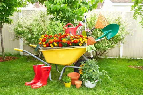 A garden scene featuring a yellow wheelbarrow filled with vibrant red and yellow flowering plants positioned on a neatly maintained green lawn. Surrounding the wheelbarrow are gardening tools and accessories, including a pair of bright red rubber boots placed on the grass, a watering can resting on the wheelbarrow, and a couple of small terracotta plant pots on the ground. To the right, there is a potted shrub growing in a white container, with colorful plastic gardening tools like a green trowel and orange rake inserted into the soil nearby. In the background, a white fence encloses the garden, with lush green trees and bushes providing a natural backdrop. The overall scene suggests active outdoor maintenance or plant care, consistent with professional gardening services such as hedge trimming available in Gipsy Hill, with natural daylight illuminating the space in fair weather conditions.