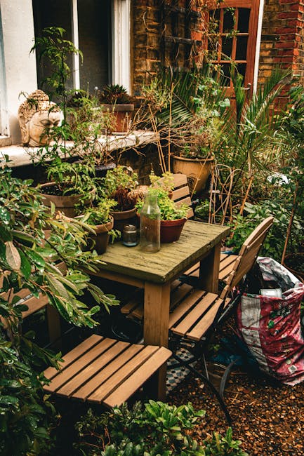 A cozy garden patio featuring a weathered wooden table surrounded by matching wooden chairs, set against a backdrop of a brick wall with a window and lush green plants. The table is decorated with var