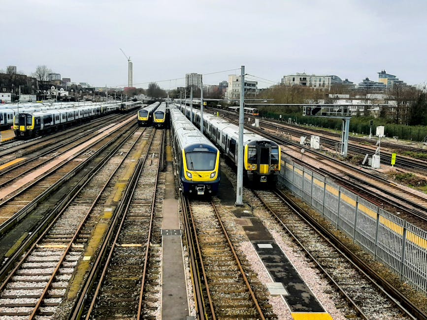A series of modern yellow and gray trains are parked on multiple parallel railway tracks in an urban setting. The trains have sleek, curved fronts with large windows, and are situated on well-maintain